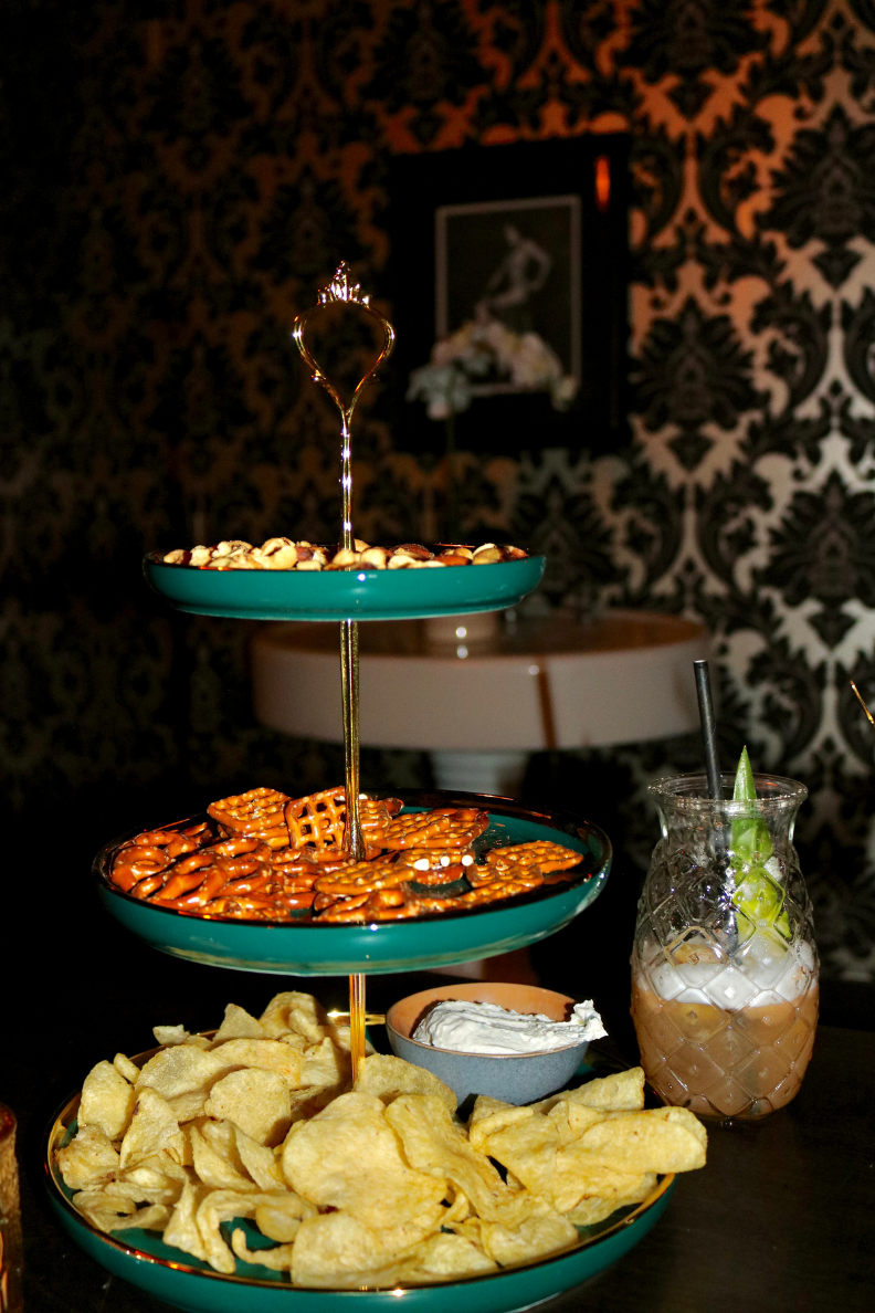 A three-tier stand of nuts, pretzels, chips and dip beside a cocktail at Society